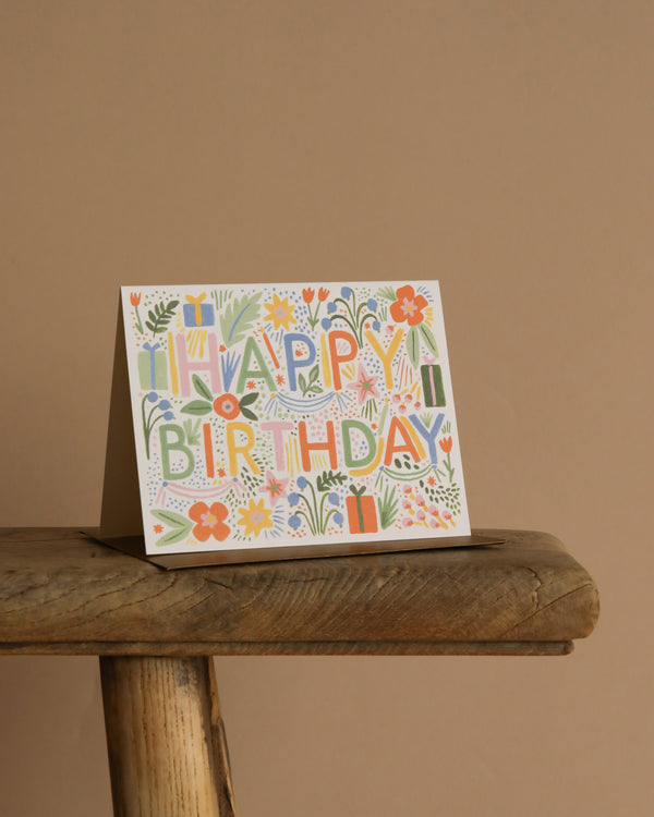 The Fiesta Birthday Card, featuring vibrant floral and gift artwork, stands on a rustic wooden stool with a metallic gold envelope against a plain beige background.