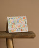 The Fiesta Birthday Card, featuring vibrant floral and gift artwork, stands on a rustic wooden stool with a metallic gold envelope against a plain beige background.