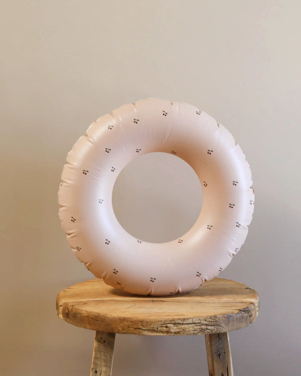 The Inflatable Junior Swim Ring - Cherry Blush in light pink, made from durable PVC with floral patterns, stands vertically on a round wooden stool against a beige background. Perfect for kids aged 3-6 years, it's ideal for summer fun.