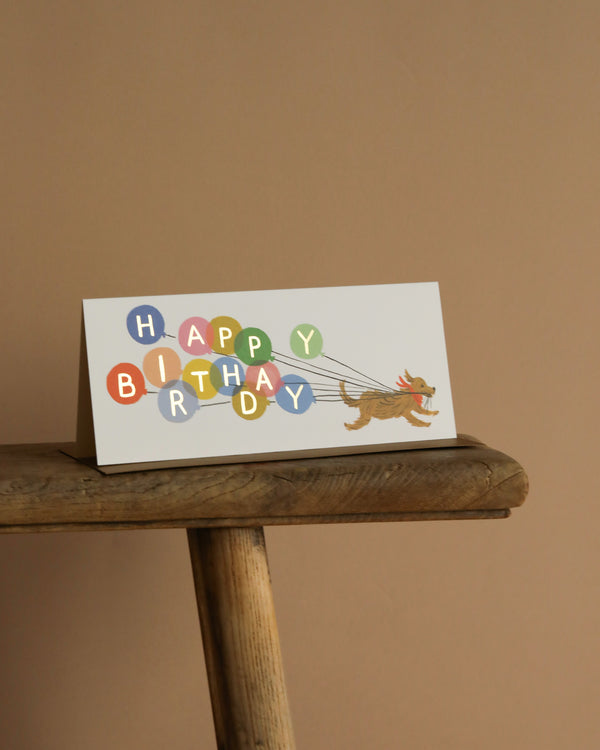 The Balloon Birthday No. 10 Card sits on a wooden table, showing a dog pulling vibrant balloons that spell "HAPPY BIRTHDAY" in gold foil against a simple beige background. Inside, the card is blank for your personal message.