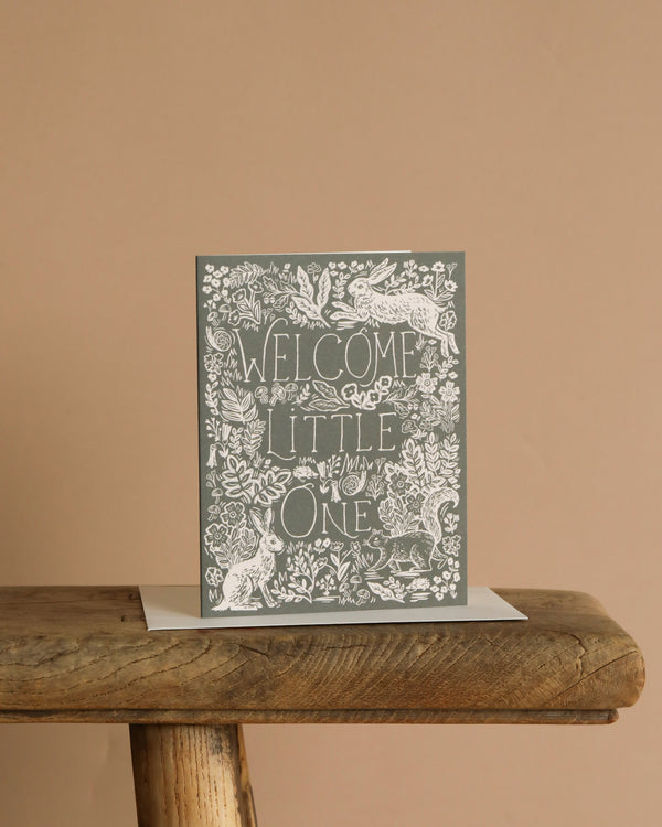 The Fable Baby Card features "Welcome Little One" in decorative white lettering on a rustic wooden table, surrounded by intricate floral and animal illustrations—perfect as a greeting card with a blank interior for your handwritten message.