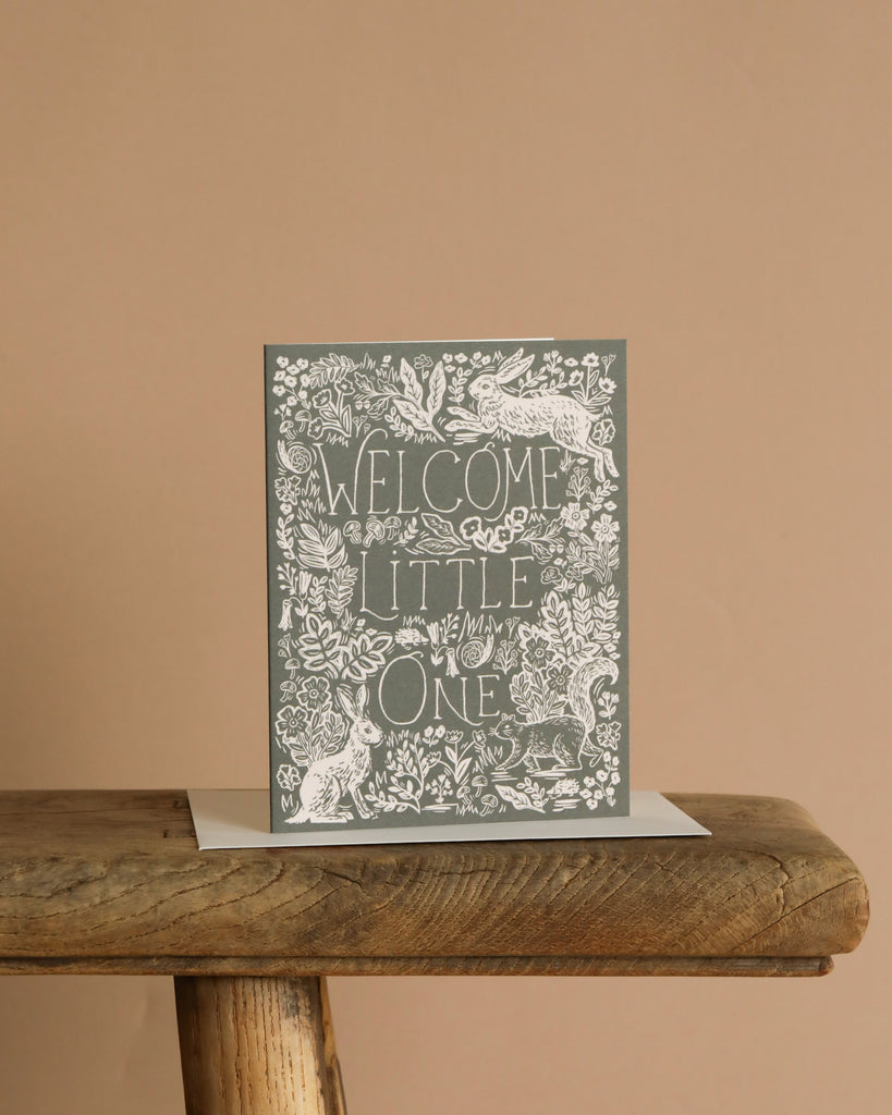 The Fable Baby Card features "Welcome Little One" in decorative white lettering on a rustic wooden table, surrounded by intricate floral and animal illustrations—perfect as a greeting card with a blank interior for your handwritten message.