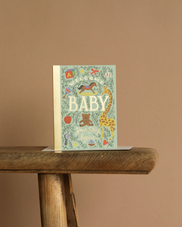 The Storybook Baby Card features colorful illustrations and metallic gold foil accents. Standing upright on a rustic wooden bench, its blank interior is perfect for your personal message.