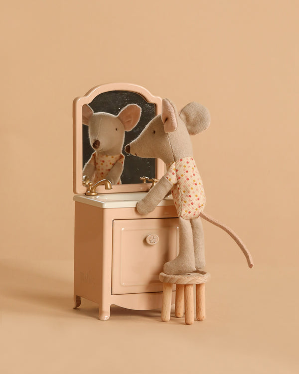 The Maileg Little Sister Mouse Vanity Set features a mouse in a floral shirt standing on a stool, gazing into a tiny mirror atop beige furniture—perfect for completing her cozy mouse house against the tan background.