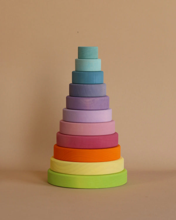 A neatly stacked pyramid of colorful wooden rings arranged by size from largest to smallest, with the largest ring at the bottom in green and the smallest at the top in light blue, all set against a plain beige background. This Pastel Stacking Tower enhances fine motor skills and eye-hand coordination.