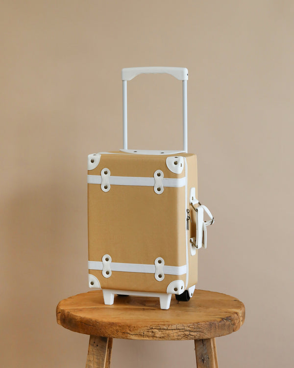 A beige Olli Ella See-Ya Suitcase - Butterscotch with white trim is perched on a wooden stool against a plain beige wall, depicting simplicity and readiness for travel.