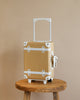 A beige Olli Ella See-Ya Suitcase - Butterscotch with white trim is perched on a wooden stool against a plain beige wall, depicting simplicity and readiness for travel.