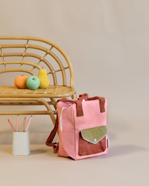 The Suzy Blush backpack from Sticky Lemon's Envelope Collection, made from recycled PET bottles with a front pocket, rests on the floor near a stool with pastel fruit decorations. A pot of colored pencils adds charm against the plain beige backdrop.