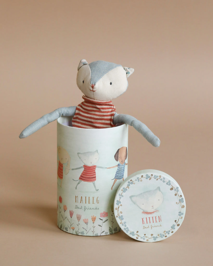 A cuddly toy, this Maileg Best Friend - Cat with a light blue head and beige body is dressed in a charming red and white striped shirt. Peeking out of a cylindrical gift box adorned with illustrations and text that reads "Maileg Best Friends" and "Kitten," it makes the perfect companion.