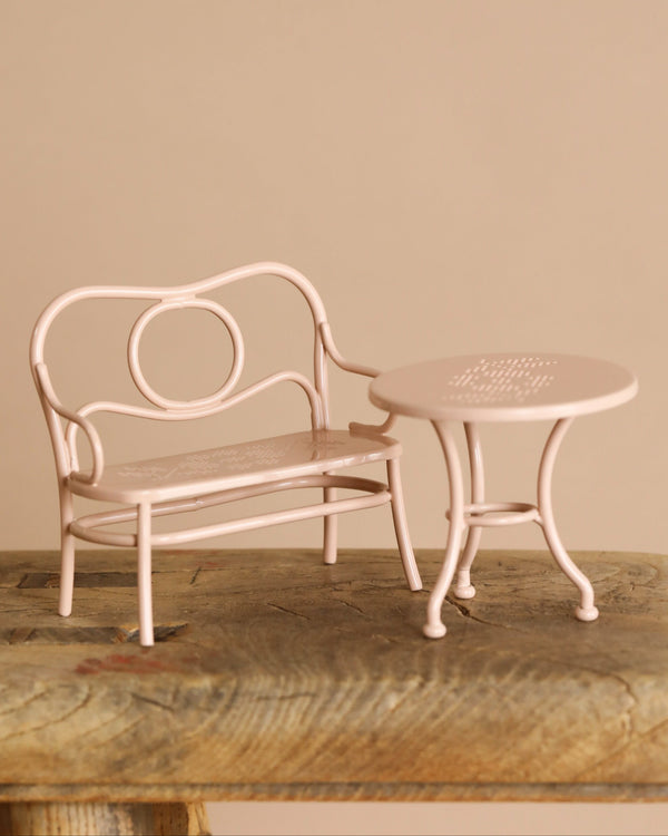 A small, pale pink metal table and chair set with intricate patterns sits on a rustic wooden surface against a plain beige backdrop. The Maileg Mouse Cafe Set furniture features curved designs, adding vintage charm ideal for an afternoon tea setting.