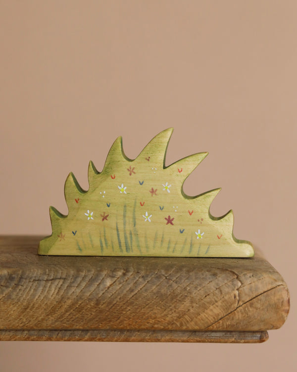 The Handmade Wooden Wildflower Patch, crafted from sustainable hardwood, features a jagged shrub design painted light green with white, red, and purple flowers. It sits on a wooden surface with a soft beige background enhanced by non-toxic plant-based stains.
