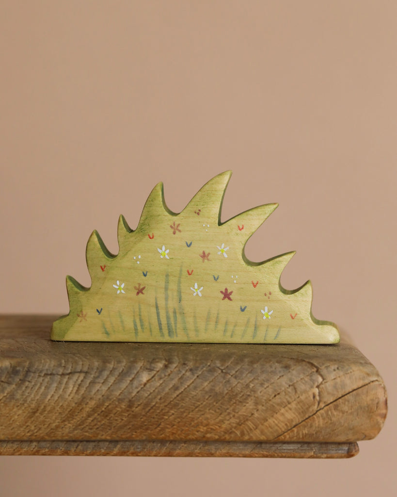 The Handmade Wooden Wildflower Patch, crafted from sustainable hardwood, features a jagged shrub design painted light green with white, red, and purple flowers. It sits on a wooden surface with a soft beige background enhanced by non-toxic plant-based stains.
