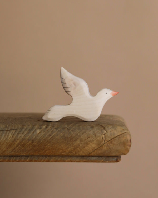 The Handmade Wooden Dove, crafted from sustainable hardwood, features outstretched wings and a pink beak, resting on the edge of a wooden surface against a beige background.