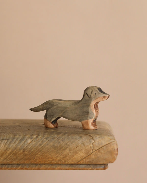 A Handmade Wooden Dachshund Dog with a natural finish stands on a rustic wooden surface. Crafted from sustainable hardwood, this unique figurine is set against a neutral beige background.