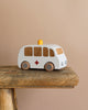 The Maileg Wooden Ambulance, crafted from solid wood, features a white body, brown windows, gray wheels, and an orange top light. It includes a red cross on the side and sits charmingly on a rustic wooden surface against a light brown background.