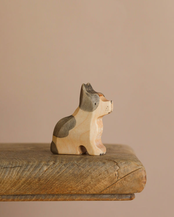 The Handmade Wooden French Bulldog, a small carved figurine with light brown and dark gray patches, finished with natural plant-based stains, sits on a rustic wooden shelf against a beige background.