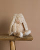 Harey the Bunny, a fluffy, cream-colored stuffed animal with a vintage look, sits on a wooden bench against a beige background. With long ears and soft, textured fur, its charm is heightened by delicate embroidered features.