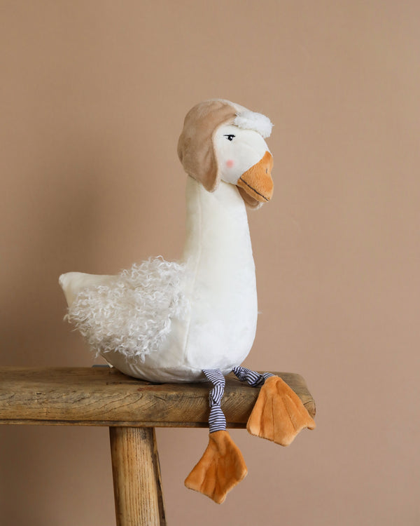 Avery the Aviator Snow Goose Stuffed Animal, made of curly polyester, sits on a wooden stool. It features a brown aviator hat with a white pom-pom, an orange beak and feet, and striped legs set against a plain beige background.