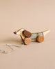 A Handmade Pull Along Dog Toy shaped like a dachshund with a checkered pattern on its body, round wheels painted with natural milk paint, and a white string against a beige background.
