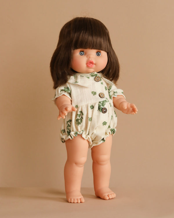 The Minikane Standing Doll (14") - Chléa wears a white romper with green leaf patterns and buttons. It has long brown hair, blue eyes, and a pinkish complexion against a beige background. With jointed arms and legs, it's anatomically correct, adding to its lifelike charm.