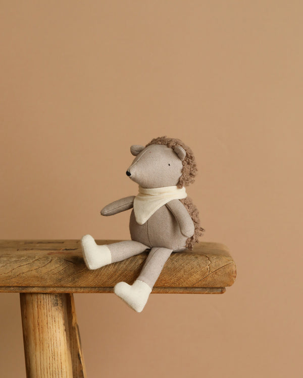Poppet The Baby Hedgehog plush, wearing a light scarf and boots, sits on a wooden table. With its pocket-sized charm against a warm beige background, it creates an inviting and cozy atmosphere as an adorable garden companion.