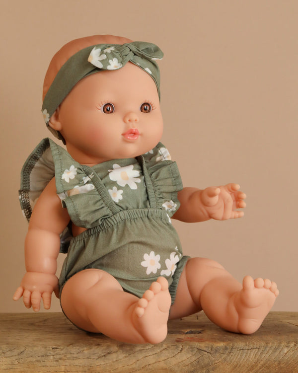 The Minikane Doll (13") - Faustine with Green Romper is displayed on a wooden surface, wearing a green outfit with white flowers and a matching headband. Made from phthalate-free vinyl, this doll encourages imaginative playtime against its beige backdrop.