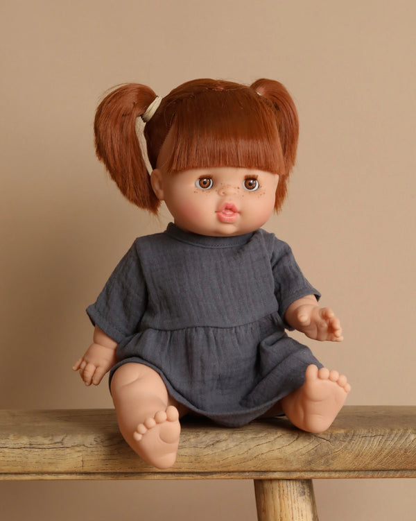 A Minikane baby doll named Raphaelle, with sleeping eyes and brown hair in pigtails, sits on a wooden bench. Wearing a dark gray dress and exuding a vanilla scent, she is set against a light beige background.