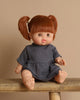 A Minikane baby doll named Raphaelle, with sleeping eyes and brown hair in pigtails, sits on a wooden bench. Wearing a dark gray dress and exuding a vanilla scent, she is set against a light beige background.