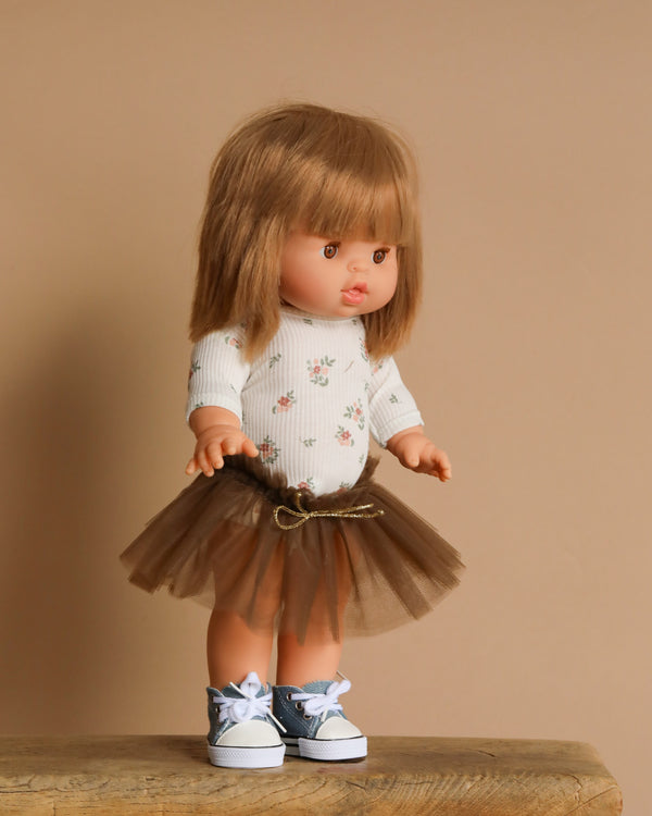 The Minikane Standing Doll (14") - Zoelie is posed on a wooden surface, dressed in a floral top, brown tulle skirt, and blue sneakers with white laces. With long brown hair and a lovely natural vanilla scent, it stands out against a plain beige background.