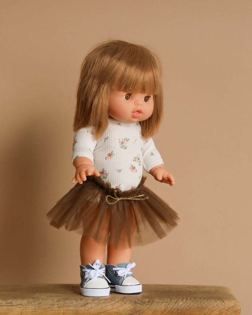 The Minikane Standing Doll (14") - Zoelie is posed on a wooden surface, dressed in a floral top, brown tulle skirt, and blue sneakers with white laces. With long brown hair and a lovely natural vanilla scent, it stands out against a plain beige background.
