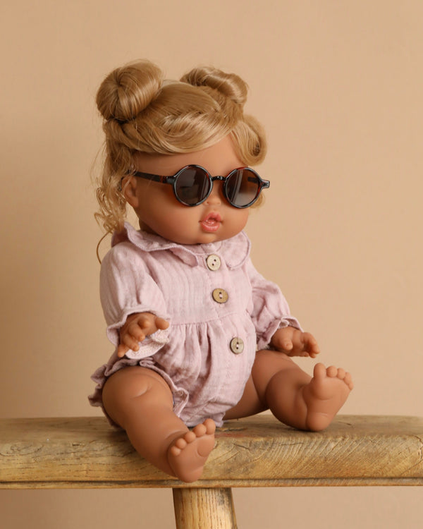 The Minikane Baby Doll (13") - Louise, featuring light brown hair in two buns, sits on a wooden bench. It wears round sunglasses and a pale pink romper with wooden buttons, standing out against a warm beige backdrop. This anatomically correct doll captures realistic details beautifully.