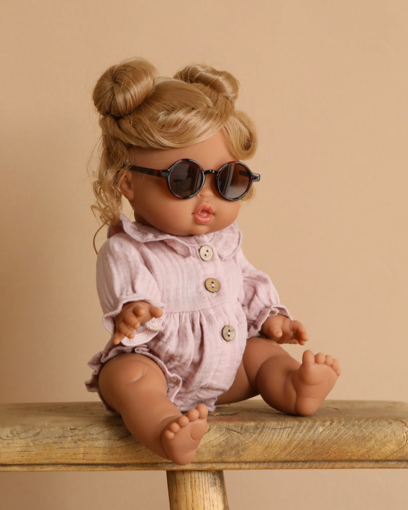 The Minikane Baby Doll (13") - Louise, featuring light brown hair in two buns, sits on a wooden bench. It wears round sunglasses and a pale pink romper with wooden buttons, standing out against a warm beige backdrop. This anatomically correct doll captures realistic details beautifully.