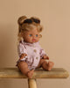 The Minikane Baby Doll (13"), Louise, with light brown hair wears sunglasses and a light pink outfit. Seated on wood against a beige backdrop, it exudes a delightful natural vanilla scent.