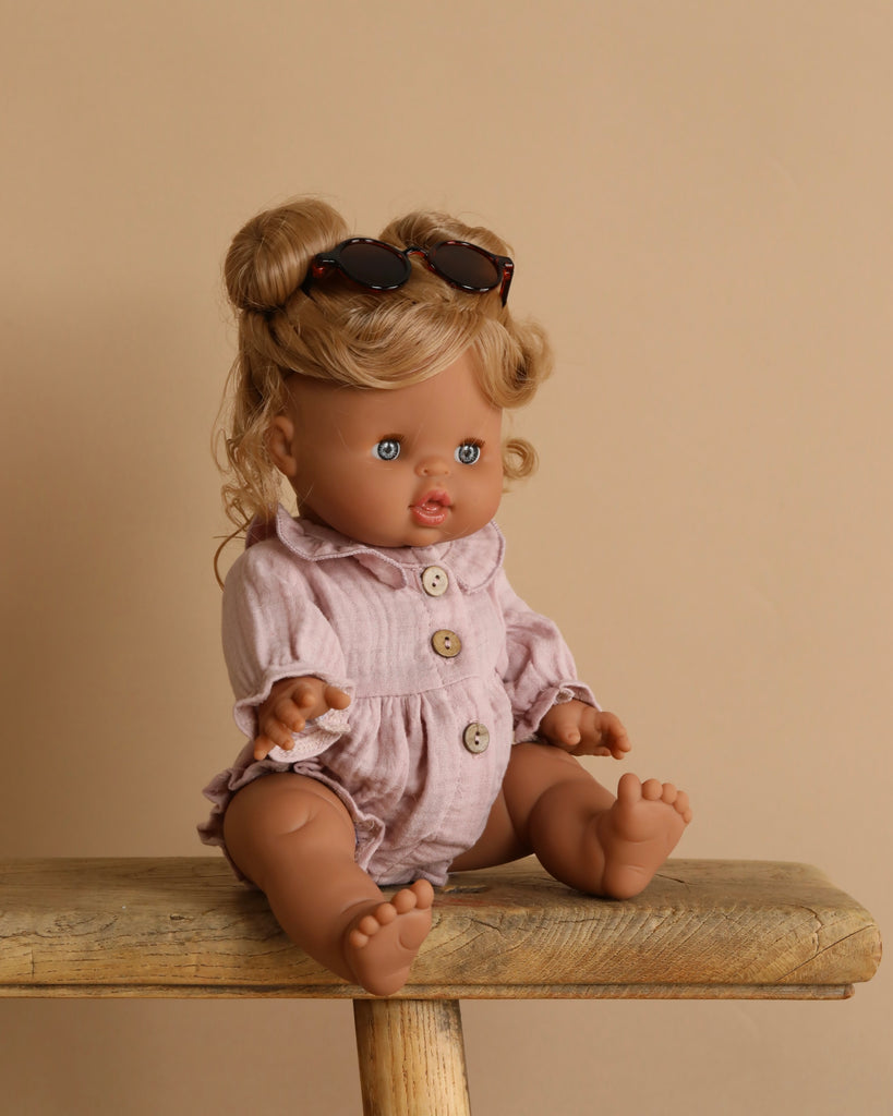 The Minikane Baby Doll (13"), Louise, with light brown hair wears sunglasses and a light pink outfit. Seated on wood against a beige backdrop, it exudes a delightful natural vanilla scent.