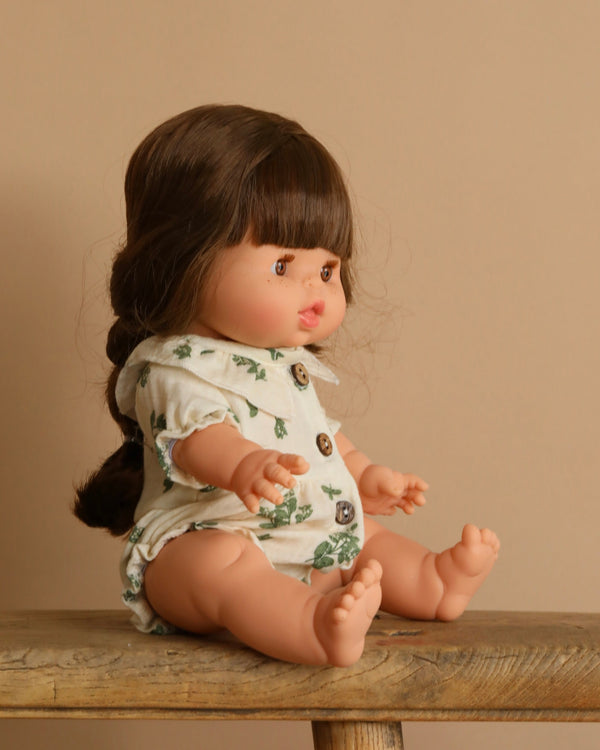 A Minikane Baby Doll (13") - Valentine is seated on a wooden surface against a beige background. With long braided brown hair, the anatomically correct doll wears a white outfit with a green floral pattern and wooden buttons, charmingly facing slightly to the left.