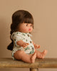 A Minikane Baby Doll (13") - Valentine is seated on a wooden surface against a beige background. With long braided brown hair, the anatomically correct doll wears a white outfit with a green floral pattern and wooden buttons, charmingly facing slightly to the left.