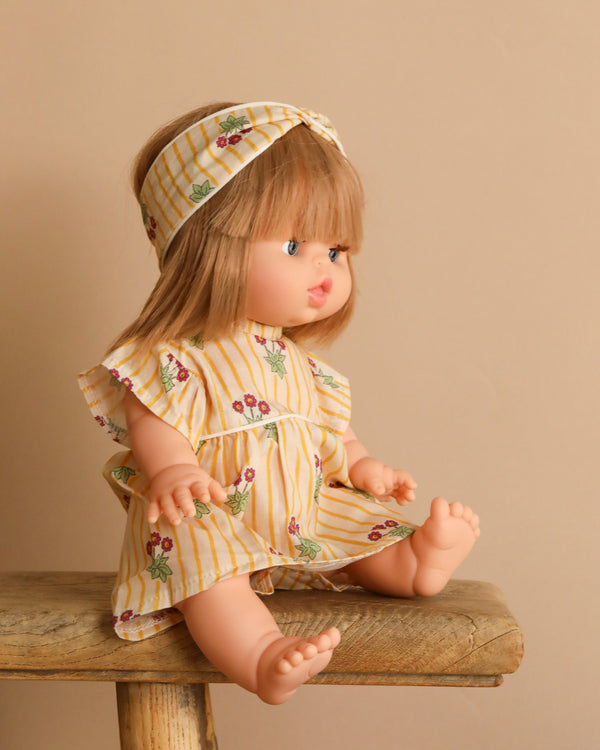 A Minikane Baby Doll With Sleeping Eyes (13") named Yzé, featuring blonde hair, sits on a wooden bench against a beige background. She wears a yellow striped dress with floral patterns and a matching headband. This adorable doll is anatomically correct for realism.