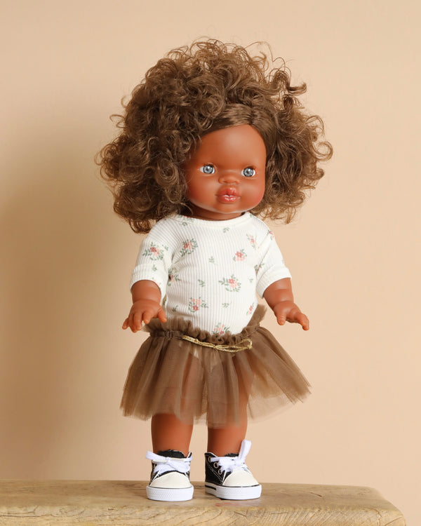 The Minikane Standing Doll (14") - Charlie, with curly brown hair, wears a floral top, brown tutu skirt, and black and white sneakers. It stands on wood against a beige backdrop and exudes a delightful vanilla scent.
