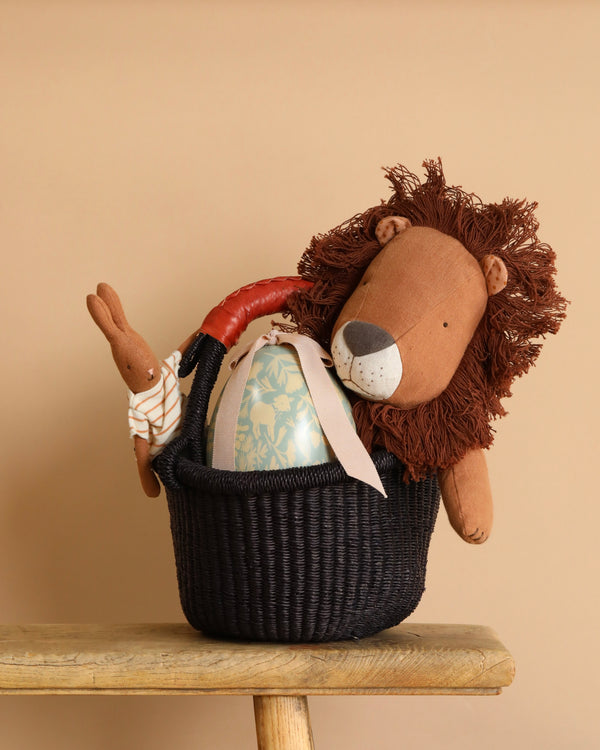 The Easter Basket Set is styled on a wooden bench, featuring a black Bolgatanga basket holding a plush lion, a fabric rabbit, and a large floral-decorated egg with a pink ribbon, all against a beige background.