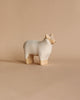 A small, minimalist ceramic sculpture resembling a handmade wooden sheep, with a light beige body and slightly darker legs and face, placed on a matching beige background.