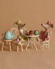 Two Maileg Ballerina Mice, with one in a yellow headband and the other in a pink hat and dress, sit at a small white table set for tea. A teal teapot, mug, cake, and macarons from the Maileg Afternoon Tea Set are on stools beside them against a beige background.