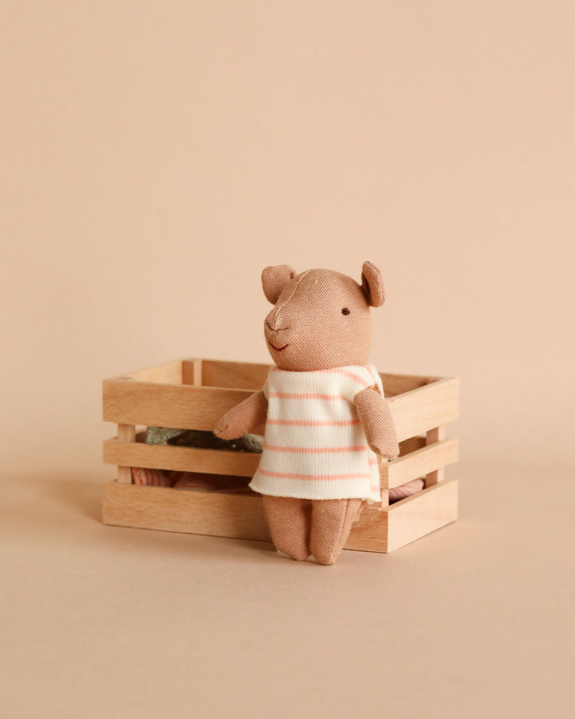 A Maileg Pig in a Box, dressed in a fairy outfit with a white dress and pink stripes, stands before a light wooden crate on a beige backdrop—ideal for a baby girl who loves pink.