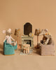 Plush toy mice are arranged in a cozy living room setting with three sitting on Maileg Soft Chairs around a tiny table, a rug in the center, and two floral-shaded lamps. A Maileg Fireplace with a mirror and clock adds charm, all part of the Maileg Living Room Set.