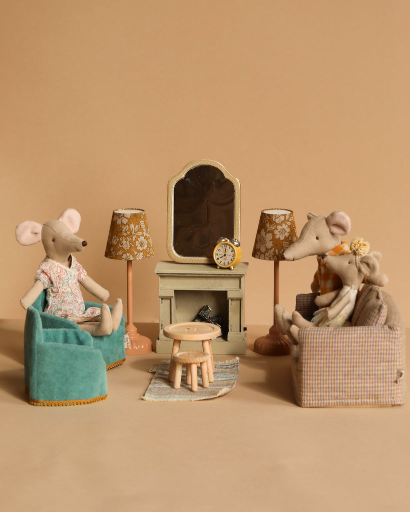 Plush toy mice are arranged in a cozy living room setting with three sitting on Maileg Soft Chairs around a tiny table, a rug in the center, and two floral-shaded lamps. A Maileg Fireplace with a mirror and clock adds charm, all part of the Maileg Living Room Set.
