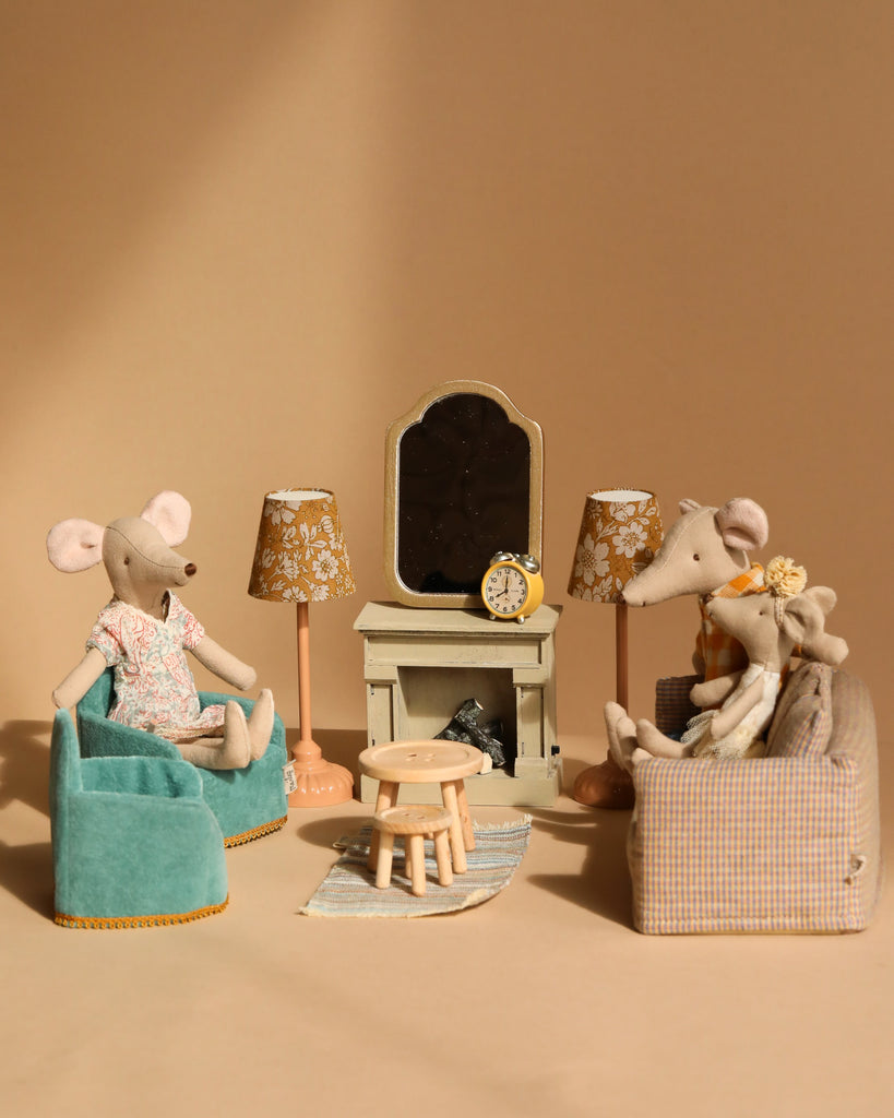 In a miniature room with the Maileg Living Room Set, two plush mice enjoy a cozy setup with a Maileg Fireplace and mirror. One relaxes on a blue chair while the other shares a plaid sofa with a smaller mouse. Two Maileg Floor Lamps flank the fireplace, and a small round table is centered.