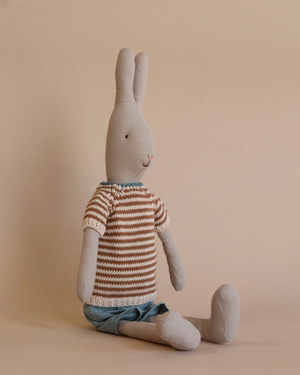The Maileg Rabbit Size 3, Classic - Knitted Shirt and Shorts is seated against a beige background. The plush gray rabbit, crafted from soft natural linen, wears a brown and white striped sweater paired with blue shorts, gazing slightly to the left. The minimalist setting accentuates the gentle and calm appearance of this timeless toy.