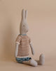 The Maileg Rabbit Size 3, Classic - Knitted Shirt and Shorts is seated against a beige background. The plush gray rabbit, crafted from soft natural linen, wears a brown and white striped sweater paired with blue shorts, gazing slightly to the left. The minimalist setting accentuates the gentle and calm appearance of this timeless toy.