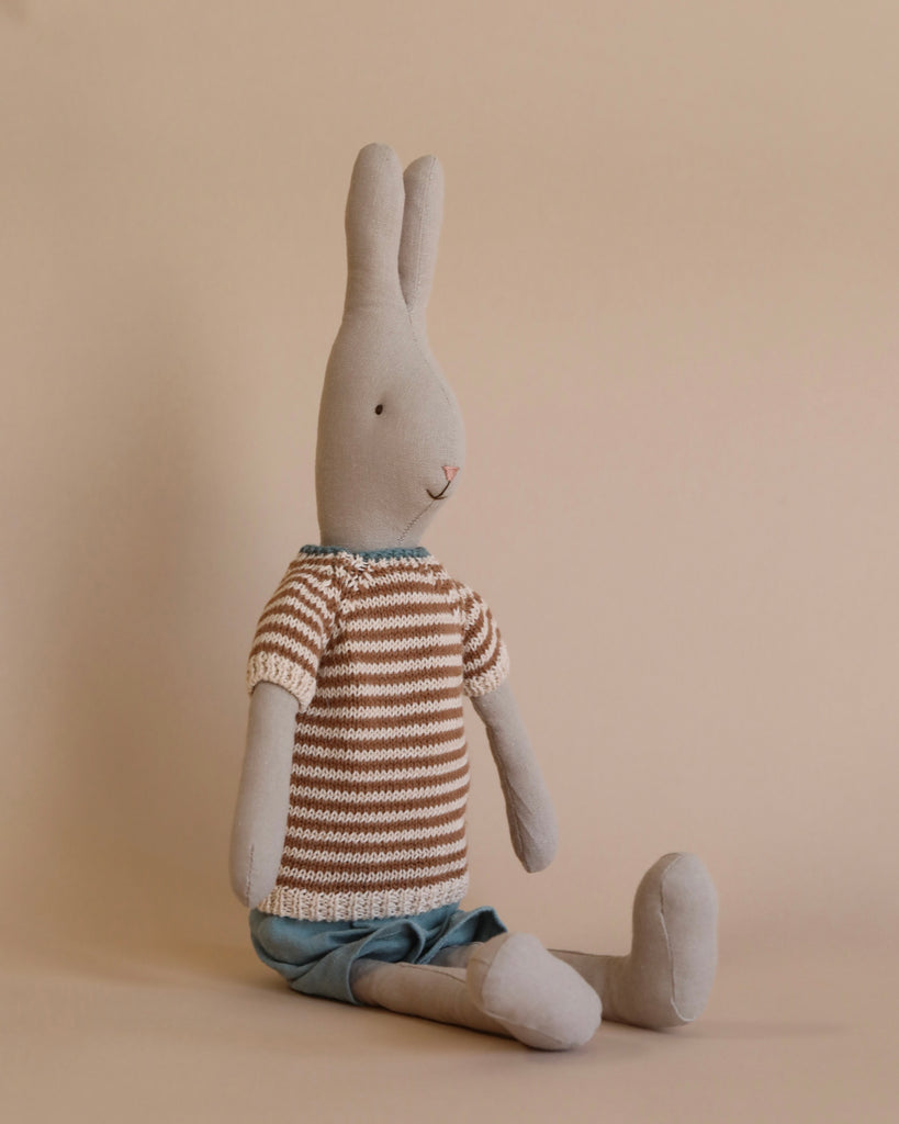 The Maileg Rabbit Size 3, Classic - Knitted Shirt and Shorts is seated against a beige background. The plush gray rabbit, crafted from soft natural linen, wears a brown and white striped sweater paired with blue shorts, gazing slightly to the left. The minimalist setting accentuates the gentle and calm appearance of this timeless toy.