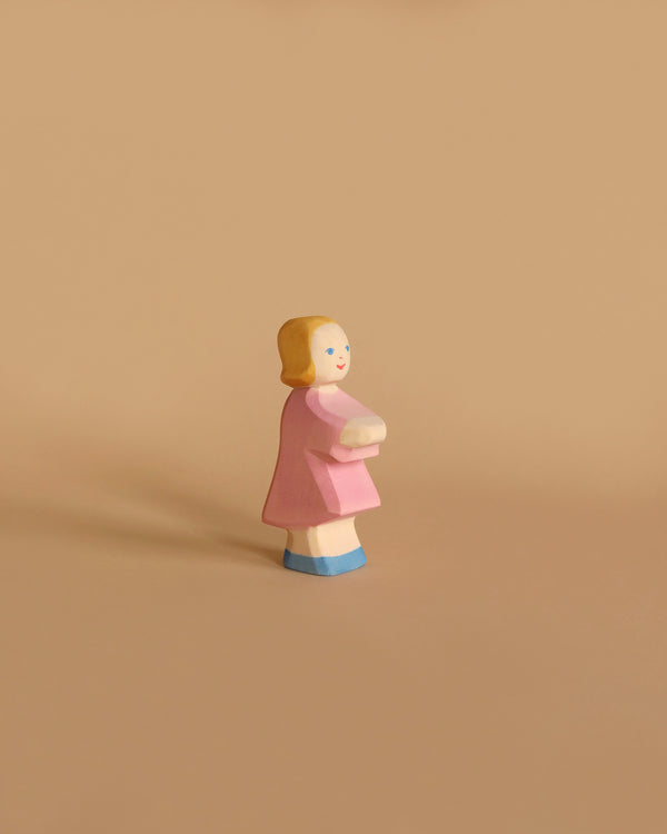 The Ostheimer Daughter in Pink Dress is a small wooden figure of a girl with short blonde hair, wearing a pink dress and blue shoes, holding a white object in front of her. Set against a plain, light brown background, this handcrafted toy from the Ostheimer brand encourages imaginative play in children.