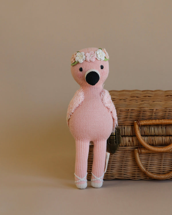 A hand knit pink Cuddle + Kind Penelope the Flamingo with a floral headband sits in front of a wicker basket on a beige background.